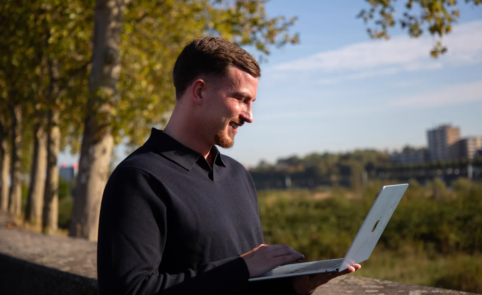 Gielke Huijs lacht terwijl hij buiten aan de Maas op zijn laptop werkt